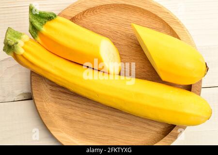 Eine ganze und eine geschnittene reife, saftige, organische, gelbe Zucchini, aus der Nähe, auf einem runden Bambustablett auf einem Holztisch, Draufsicht. Stockfoto