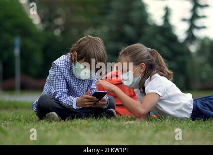 Die Kinder gehen zurück zur Schule. Niedliche Schüler mit Rucksäcken. Junge und Mädchen in Sicherheitsmasken liegen auf dem Gras Stockfoto