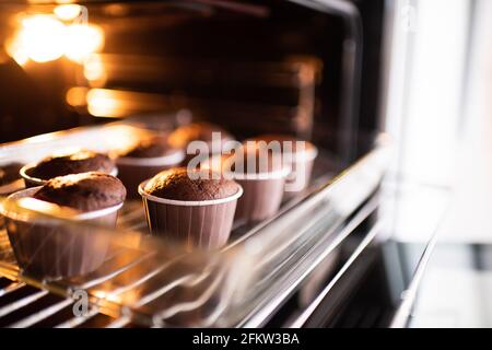 Öffnen Sie den Ofen mit leckeren Schokoladenmuffins in Backform auf dem Tablett zu Hause. Frische Cupcakes im Ofen aus nächster Nähe backen. Stockfoto