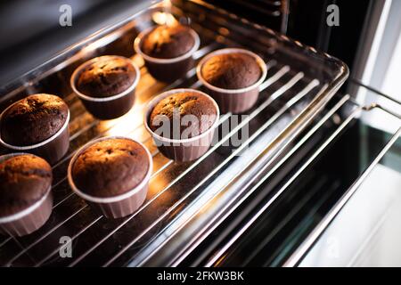 Öffnen Sie den Ofen mit leckeren Schokoladenmuffins in Backform auf dem Tablett zu Hause. Frische Cupcakes im Ofen aus nächster Nähe backen. Stockfoto