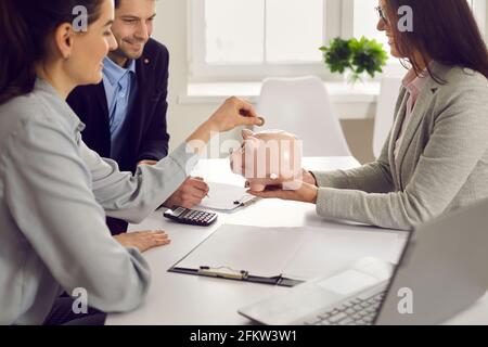 Seitenansicht eines Ehepaares, das eine Münze in eine Sparschwein legt, die in den Händen eines Bankmanagers gehalten wird. Stockfoto