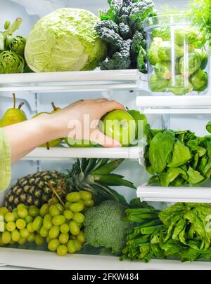 Frisches grünes Gemüse und Obst im Kühlschrank. Die Frau nimmt den grünen Apfel aus dem offenen Kühlschrank. Gesunde Ernährung. Stockfoto