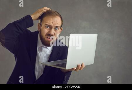 Verwirrt Geschäftsmann hält einen Laptop und kratzt seinen Kopf, während er auf einem grauen Hintergrund steht. Stockfoto