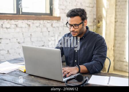 Fokussierter hindu-Freiberufler, der Brillen mit Laptop im modernen Bürobereich trägt, konzentrierter Mann mit gemischter Rasse, der tippt, Messaging, indischer Programmierer entwickelt Software, schaut auf den Computerbildschirm Stockfoto