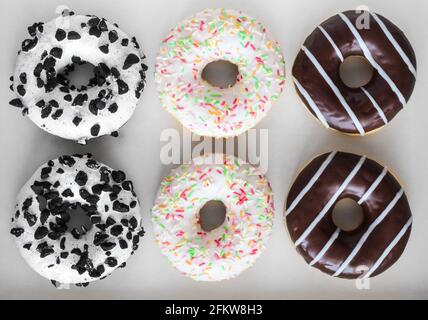 Flaches Lay-Bild von sechs Ringdonuts mit weißer Glasur und bunten Hunderttausenden, Schokolade und Streifen und weißer Glasur mit schwarzen Keksen Stockfoto