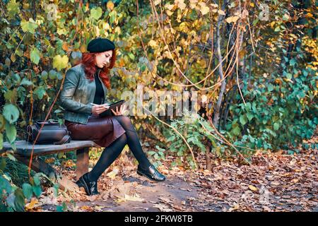 Frau mit grafischem Tablet auf Parkbank. Herbstlandschaft im Hintergrund. Rothaariges Mädchen in einer Baskenmütze und einer modischen Jacke mit Lederrucksack. Designer oder Freiberufler in der Natur. Stockfoto