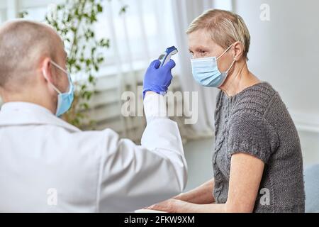 Ein Arzt misst die Temperatur einer älteren Frau in ihrem Zuhause mit einer Schutzmaske. Coronovirus-Infektionsdiagnosekonzept Stockfoto