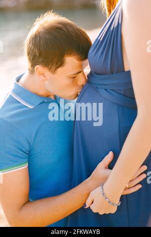 Mann küsst schwanger Bauch der Frau in langen blauen Kleid An einem sonnigen Tag Stockfoto