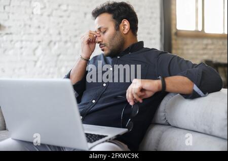 Müder, gemischter hinduistischer Unternehmer zieht die Brille ab und fühlt die Augenbelastung durch die Online-Arbeit mit dem Laptop, der indische Mann sitzt auf der Couch und massiert die Augen, fühlt Kopfschmerzen und Mangel an Energie Stockfoto