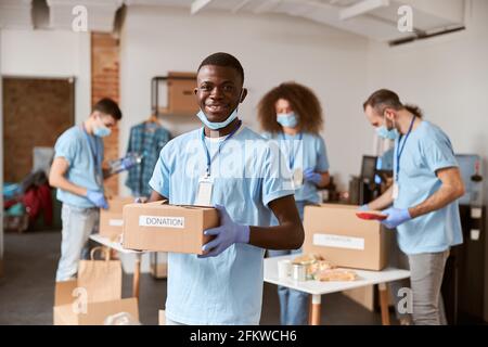 Porträt eines jungen freiwilligen aus afrika, in blauer Uniform, Schutzmaske und Handschuhen, lächelnd und mit Pappkarton zur Spende Stockfoto