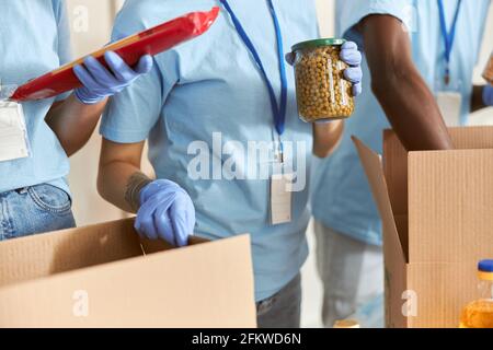 Nahaufnahme eines Freiwilligen in Schutzhandschuhen, der ein Glas Erbsen hält, während er Lebensmittel in einem Karton sortiert und verpackt, um an einem Spendenprojekt zu arbeiten Stockfoto