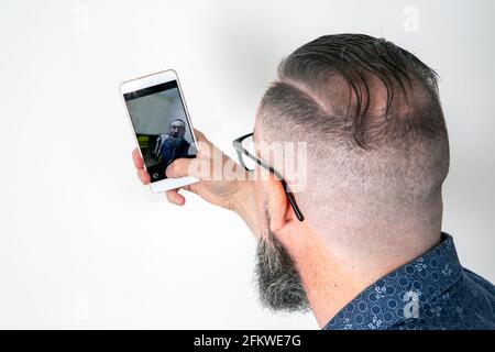 Bärtiger Hipster, der sein Smartphone benutzte, um ein Selfie zu machen, von hinten gesehen und auf dem Handy reflektiert Stockfoto