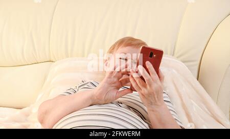 Eine ältere Frau liegt mit einem Telefon im Schlafzimmer In ihren Händen Stockfoto