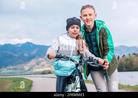 Bruder hilft der Schwester und lehrt erste Schritte im Reiten. Kinder auf einem Radweg am Flussufer. Happy Kindheitskonzept Bild. Stockfoto