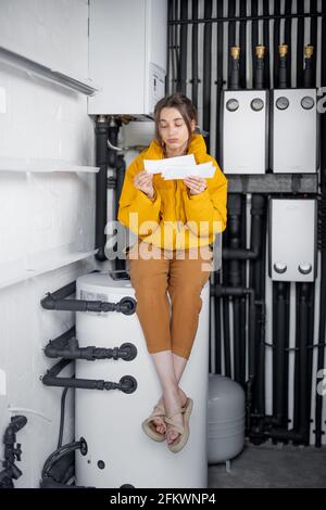 Verwirrt betrachtet eine Frau die Stromrechnungen und vergleicht die Daten mit dem Messgerät in der elektrischen Zentrale zu Hause. Konzept der Haushaltsausgaben Stockfoto