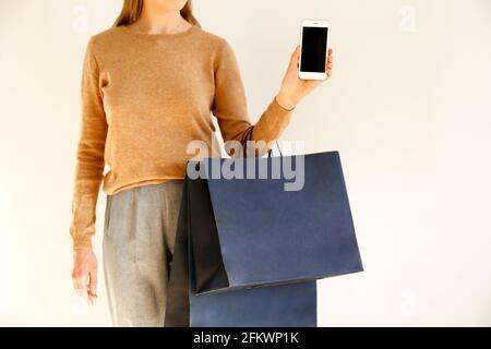 Junge schlanke Frau im klassischen modischen Outfit, brauner Pullover und lockere graue Hosen gingen einkaufen, hielten Papiertaschen und zeigten einen leeren Bildschirm smartpho Stockfoto