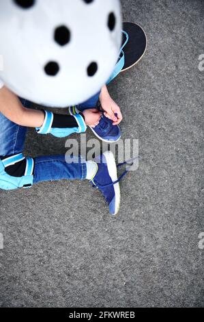 High-Angle-Ansicht des nicht erkennbaren Jungen in Schutzhelm für Skateboard Binde Schnürsenkel sitzen auf Asphalt des Spielplatzes Stockfoto