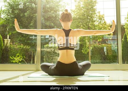 Attraktive junge Yogi-Frau in schwarzer Sportkleidung, die Yoga praktiziert, in Padmasana-Lotus-Pose sitzt und sich in einem geräumigen Raum mit durchgehendem Wind dehnt Stockfoto