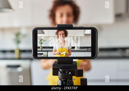 Frau youtuber nimmt sich mit ihrem Smartphone in ihrer Küche auf, während sie ein Salatrezept zubereitet, es gibt natürliches Licht und sie trägt ein gelbes A Stockfoto