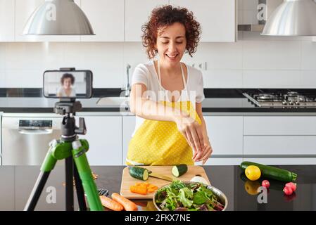 Frau youtuber nimmt sich mit ihrem Smartphone in ihrer Küche auf, während sie ein Salatrezept zubereitet, es gibt natürliches Licht und sie trägt ein gelbes A Stockfoto