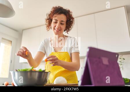 Junge kaukasische Frau mit lockigen Haaren bereitet in der Küche ihres Hauses einen Salat zu, während sie ihr digitales Tablet betrachtet, es ist Tageslicht und es gibt na Stockfoto