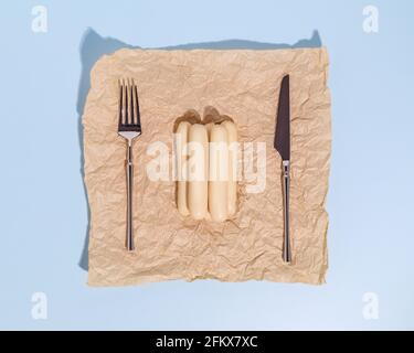 Horizontale Lebensmittelzusammensetzung mit Geflügelwürsten auf Packpapier mit Messer und Gabel auf blauem Hintergrund. Gesunde, Nahrungsergänzungsmittel Fleisch prod Stockfoto
