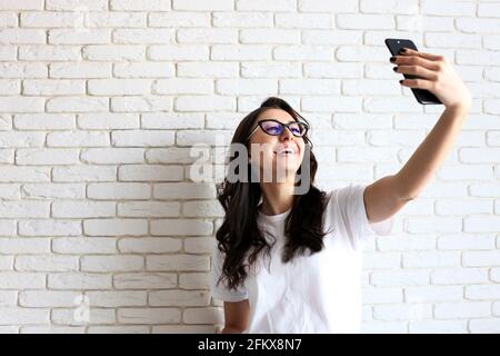 Schöne junge Frau in Katzenaugenbrille, Selfie auf mobilen Gadget-Gerät zu machen. Hipster Frau trägt Mama Jeans & einfarbiger weißer Hemd fotografieren Stockfoto