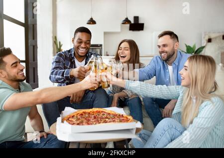 Unbeschwerte Gruppe von glücklichen Freunden, die Party genießen, Pizza essen, mit Bierflaschen toasten, etwas zu Hause feiern Stockfoto