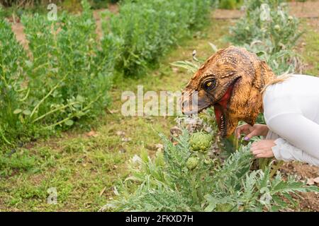 Frau mit Dinosaurier Tier Kopf Maske essen Artischocken im Gemüsegarten. Stockfoto