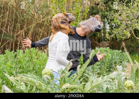 Frau mit Dinosaurier Kopf Maske angreifende Mann mit Pferdekopf im Gemüsegarten, im Freien in der Natur. Stockfoto