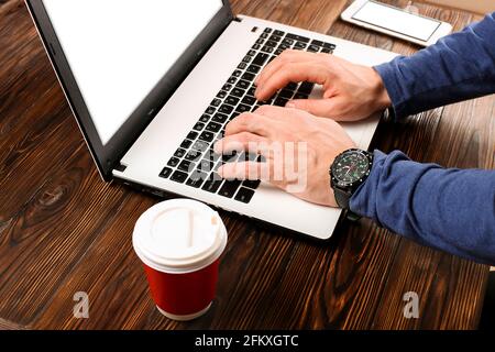 Nahaufnahme der männlichen Hände mit Armbanduhr, die auf der tastatur des pc-Laptops tippt. Roter Einweg-Papierbecher mit Plastikkappe aus dem Café auf Holz Stockfoto