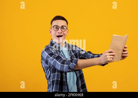 Überraschte den männlichen Studenten, der Lehrbuch in Armdistanz hielt und die Kamera schockiert auf orangenen Studiorugen ansah Stockfoto