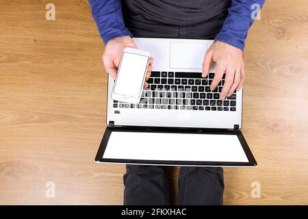 Draufsicht, junger Mann mit pc-Laptop-Computer und Handy auf dem Schoß, auf Holzboden im Hintergrund sitzend. Aufnahme von männlichen Händen auf der Tastatur, holdi Stockfoto