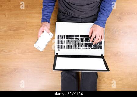 Draufsicht, junger Mann mit pc-Laptop-Computer und Handy auf dem Schoß, auf Holzboden im Hintergrund sitzend. Aufnahme von männlichen Händen auf der Tastatur, holdi Stockfoto