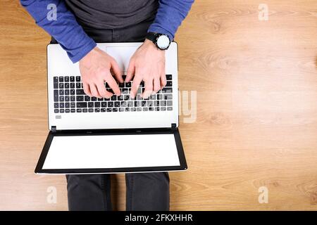 Draufsicht, junger Mann mit pc-Laptop-Computer und Handy auf dem Schoß, auf Holzboden im Hintergrund sitzend. Aufnahme von männlichen Händen auf der Tastatur, holdi Stockfoto