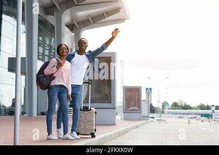 Glückliches Junges Schwarzes Paar, Das Mit Koffer In Der Nähe Des Flughafenterminals Steht Und Ein Taxi Nimmt Stockfoto
