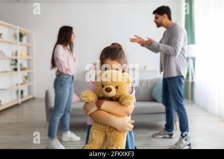 Kinder und Familienprobleme Konzept. Kleines Mädchen, das sich hinter einem Teddybären versteckt und unter den Streitigkeiten der Eltern leidet Stockfoto