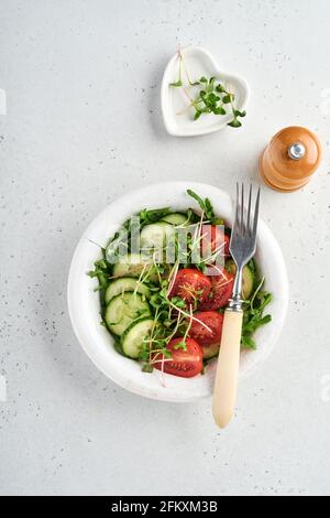 Frischer Salat mit Tomaten, Gurken, Gemüse, mikrogrünen Radieschen auf weißem Teller auf grauem Steingrund. Blick von oben. Konzept vegan und gesund Stockfoto