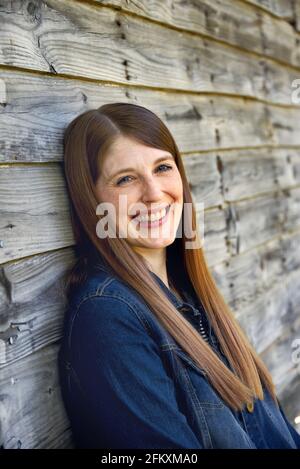 Ein großes Lächeln erhellt das Gesicht einer jungen Frau, die sich an ein Holzgebäude lehnt. Sie trägt eine blaue Jeansjacke. Stockfoto