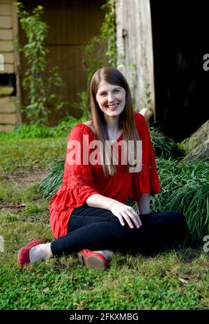 Auf dem Gras sitzend, posiert die junge Frau in schwarzen Hosen, rotem Hemd und Schuhen vor der Holzscheune. Stockfoto