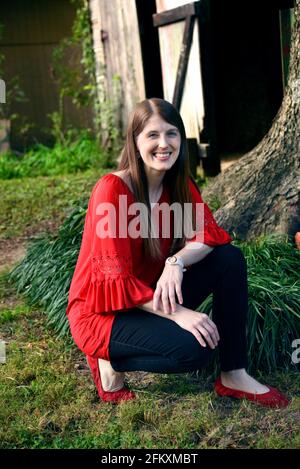 Die junge Frau kniet in schwarzen Hosen und rotem Hemd auf dem Hof. Sie kniet neben einem Baum und einer rustikalen, hölzernen Scheune. Sie lächelt und ist glücklich. Stockfoto