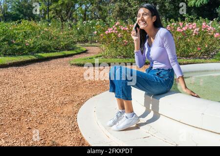 Schöne junge Latina, die am Morgen lächelnd auf dem Rand eines Springbrunnens sitzt und am Handy telefoniert. Stockfoto