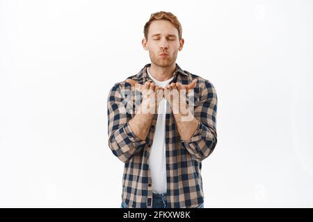 Leidenschaftlicher Rotschopf-Mann, der einen Kuss schickt, auf offene Hände bläst, als er Mwah auf die Kamera bläst und romantisch vor weißem Hintergrund steht Stockfoto