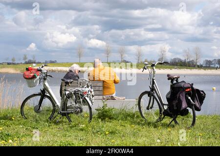 Ein älteres Rentnerpaar sitzt auf einer Steinbank und genießt den Blick auf einen See und die Landschaft. Die Elektrofahrräder sind im Gras geparkt Stockfoto