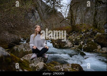 Mädchen sitzt auf einem großen Stein und meditiert in der Nähe eines Wasserfalls neben einem Bergfluss. Stockfoto