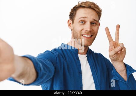 Sagen Sie Käse. Lächelnder, hübscher Rotschopf-Mann, der Selfie mit ausgestreckter Hand hält, ein Peace V-Zeichen zeigt und glücklich aussieht, über Weiß stehend Stockfoto