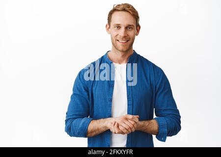 Wie kann ich Ihnen helfen? Lächelnder, freundlicher und höflicher Rotschopf-Mann, der zufrieden und bereit steht, die Hände in der Nähe der Brust hält, seinen Service anbietet, Kunden unterstützt Stockfoto