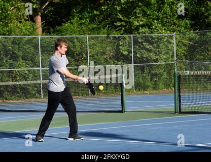 Junger Mann schwingt und verbindet sich während eines Spiels mit Pickle Ball. Er hat eine schwarze Trainingshose und ein graues Hemd. Er spielt auf einem Tennisplatz im Freien. Stockfoto