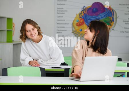 Zwei Mädchen sitzen auf Schreibtischen mit Laptop und Notebook und studieren in modernen High School-Gebäuden, glücklich gebildete weibliche Teenager Freunde Stockfoto