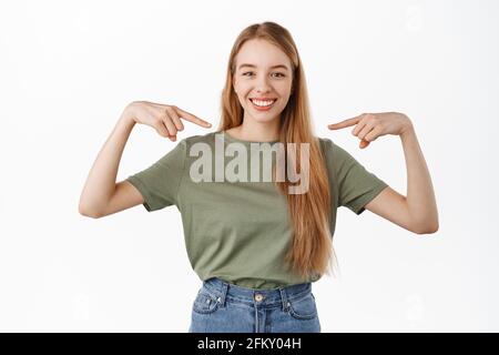 Selbstbewusstes blondes Mädchen lächelt mit weißen perfekten Zähnen, zeigt auf sich selbst und schaut entschlossen auf die Kamera, selbstfördernd, ehrenamtlich, Pick me Stockfoto